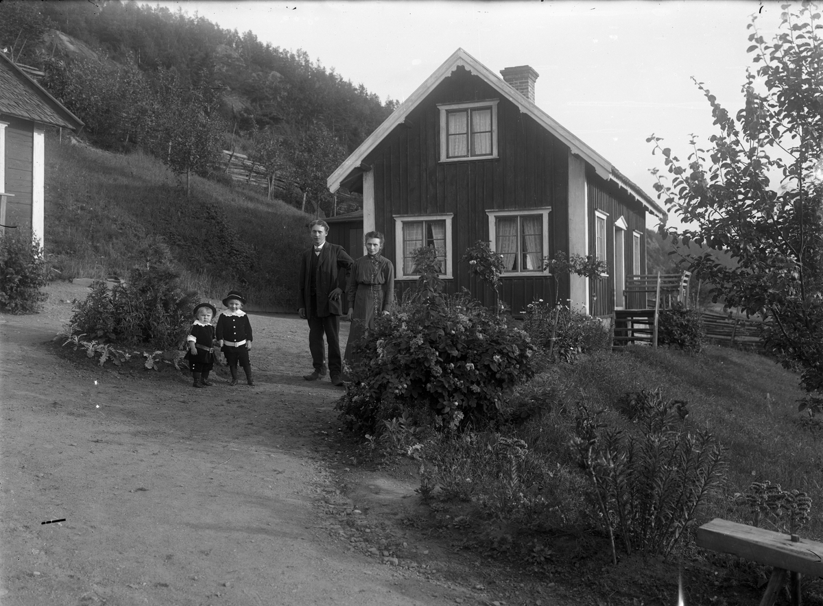 En man och en kvinna samt två små barn står på en gårdsplan framför gaveln på ett bostadshus med locklistpanel, en våning och inredd vind. Till vänster skymtar en ekonomibyggnad. Tomten lutar kraftig från berget till vänster i bild.
