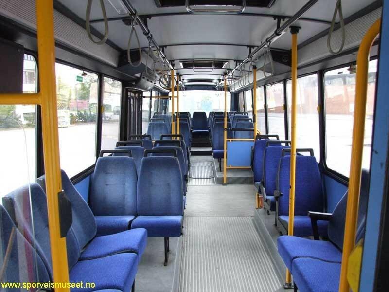 Rød buss med blå dører og et blått bånd på langsiden. Bussen har to dobbeltdører. Bussens interiør består av blå seter, gule stenger og gråe tak og gulv. 