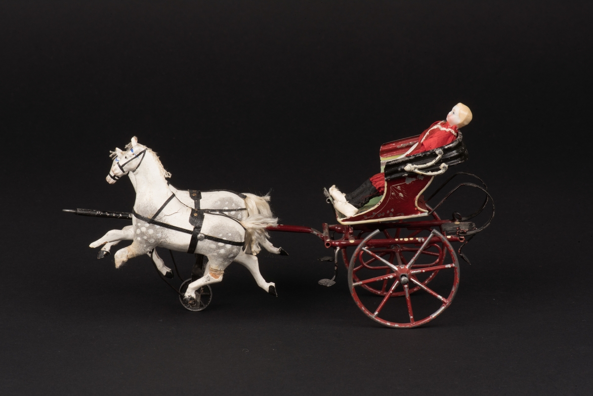 Leksaksekipage som består av en vagn av plåt, dragen av två hästar av trä.
Vagnen är tillverkad av plåt och målad i rött, grönt och svart. Vagnen är öppen med nedfälld sufflett, ensitsig och med två hjul.
På sittplatsen finns en nedböjd metallten där den ursprungliga dockan sattes.
Vagnen är förspänd med två apelkastade hästar, vita och grå med fläckar. Hästarna är av trä överdragna med en massa. Man och svansar är av silke, seldonen av läder.
Mellan hästarna finns en hjuldriven funktion som gör att hästarna rör sig upp och ned och ser ut att trava när man skjuter ekipaget framåt.
