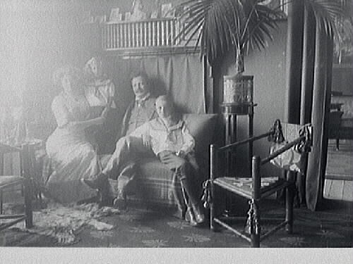 Interiörbild av familjen Hellborg som sitter på en soffa; makar med två barn. Intill står en tidstypisk "munkstol" och palm på peidestal.