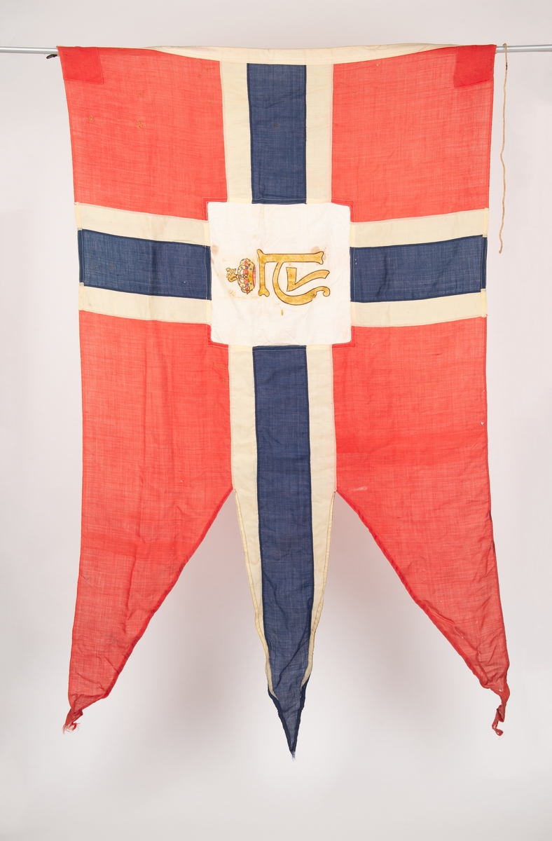Norsk splittflagg med tunge. Hvit kvadratisk felt i midten med kong Haakon VII kronede monogram i gull og brunt. Tau i innerkanten med messingkrok. Diverse stoppede småhull.