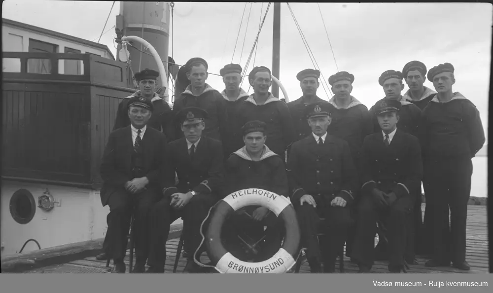 Gruppebilde av mannskap fra dampbåten "HEILHORN" fra Brønnøysund, ikledt  ulike sjøuniformer. Det er tatt  på en trekai hvor båten ligger inntil. Bildet har tilhørt Lars Rygh fra Hønefoss, som var med på mineryddings arbeid i Varangerfjorden 1917 - 1918.
