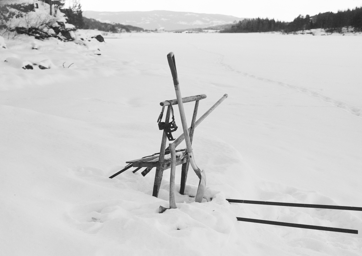 Sparkstøttingen til yrkersfiskeren Paul Stensæter (1900-1982) fra Hole kommune i Buskerud, fotografert vinteren 1973. Dette bildet er tatt på den snødekte isen på Steinsjorden, en sidearm til Tyrifjorden på Ringerike. Her drev Stensæter næringsfiske nesten hele året. Vinterstid fisket han stort sett med garn, dels i åpent vann lengre ute i Tyrifjorden, dels under isen her på Steinsfjorden. Det utstyret som lå på sparken var knyttet til det sistnevnte fisket. Garna ble satt og trukket ned i vannet via hull som Stensæter hadde hogd i isflata. Garna ble trukket med to døgns mellomrom. I løpet av en slik periode dannet det seg ofte isskorper i slike hull. Denne isen ble hogd i stykker ved hjelp av isbila som sto støttet mot sparken. Sannsynligvis gikk det også an å bruke øks i slikt arbeid. Den lette aluminiumsspaden som sto reist mot sparken ble brukt til å fjerne issørpe og snø fra hullene før garna kunne trekkes. På styret på sparken hang det også brodder som kunne festes under skosålene til fiskeren. Disse kunne være nyttige tidlig på sesongen, da Stensæter måtte gå på hard blankis når han skulle trekke garna sine. Når det var kommet snø i slike mengder som dette fotografiet viser gav broddene neppe bedre feste. I bakgrunnen, på den snødekte isflata, skimter vi fotsporene etter Steinsæter, som altså med to døgns mellomrom gikk ut til de stedene der han hadde satt garn.

I 1970-åra var etnologen Åsmund Eknæs fra Norsk Skogbruksmuseum flere ganger på besøk hos Paul Stensæter for å observere ham i aktivitet som fisker og intervjue ham om denne virksomheten. Eknæs oppsummerte det han fikk se og høre om garnfisket under isen på Steinsfjorden slik:

«Sikfiske med garn. Dette fisket foregikk på to steder, i åpent vann ute i Tyrifjorden og under isen i Steinsfjorden. Tyrifjorden er ofte åpen langt utover vinteren og det hender at den ikke legger seg i det hele tatt. De dro da i båt over fra Steinsfjorden og satte garn på ganske store dyp, 20-40 favner. Når det ble fisket for fullt ble det brukt 20 garn. 10 sto ute mens de øvrige var hjemme til tørking.

Garnfisket under isen i Steinsfjorden begynner så fort isen legger seg om høsten, fordi tynn og gjennomsiktig is er en fordel når garna skal settes ut første gang. Å sette ut garn under isen kaller Paul for øvrig «å høgge ut garna». Dette krever en spesiell teknikk. Er isen gjennomsiktig, foregår det på følgende måte: To hull hogges med så lang avstand som lengda på garnet. Ei rett granstang på 7-8 m stikkes ned i det ene hullet med den tynneste enden først. I den tykkeste enden er det et hull hvor det blir festet ei snor. Stanga blir nå skjøvet i full fart mot det andre hullet. En viktig detalj ved denne stanga er at den skal være nyhogget. Da ligger den dypere i vannet og skubber mindre mot isen. Stanga går ikke helt fram til det andre hullet. Der den stopper blir det hogget et mindre hull og den skyves videre ved hjelp av en kjepp med ei kløft i enden. Når snora er brakt fram på denne måten, er det en enkel sak å trekke garnet under isen. 

Men er isen ugjennomsiktig, slik at det er umulig å se stanga, må Paul gjøre det på en annen måte. Da finner han ei lang stang med god krumming på. Så hogger han hull så tett at stanga kan stikkes ned i det ene og komme opp igjen gjennom det neste.

Garnet er nå på plass under isen og står på bunnen på 5-10 favners dyp. Fra hver ende av garnet går det ei tynn snor opp til hullet i isen. Snorene går ikke opp gjennom hullet, men er festet litt ved siden. Dette er gjort for å hindre at han hogger dem av når is som har dannet seg i hullet skal fjernes.

Dagens nylongarn tåler å stå ute hele vinteren. Tidligere, da lin og bomull var mest brukt, var det nødvendig å ta garna opp og tørke dem. Av de 20-40 garn som Paul brukte, var halvparten til tørk.

Ettersynet, som gjerne foregår annenhver dag, begynner med at han får tak i snorene fra garnet ved å stikke en pinne med krok på innunder isen. I den borteste enden løsner han garnsnora og fester isteden ei lang nylonsnor til garnet. Tidligere brukte han snor av tvunnet hestetagl. Snora er så lang at den også rekker bort til der han står oppå isen. Ved trekkinga tar han ut fisken etter hvert. Når hele garnet er trukket, blir han stående på samme sted, tar tak i snora og drar garnet ut igjen samtidig som han passer på at det går ordentlig.

I sterk kulde vil det våte garnet fryse til en klump og være umulig å sette igjen. For å hindre dette hogger han ei grop i isen bak hullet og fyller denne med vann. Etter hvert som han trekker garnet putter ha det ned i gropa og det holder seg opptint til det skal settes igjen.

Når det blir mildvær og fare for at isen skal gå opp og komme i drift, må det tas spesielle forholdsregler for å hindre at garna blir dratt med isflak og forsvinner. Paul binder i slike tilfelle inn en bit snelletråd i den snora som går fra isen og ned til garnet. Kommer isen i drift, vil snelletråden ryke, og garnet blir stående igjen på samme sted. Det er da en forholdsvis enkel sak å sokne etter det.

Garnfisket ga jevne tilførsler av sik gjennom det meste av vinteren. Vintersiken i Steinsfjorden er noe mindre enn høstsiken. Det går omkring 4 på kiloen, mot 3 om høsten. Fangstene varierer fra noen få til 25-30 pr. garn. Med 10 garn ute vil han kunne få opptil 200-300 sik eller 50-70 kilo. Men da blir garnene trukket bare annenhver dag.»
