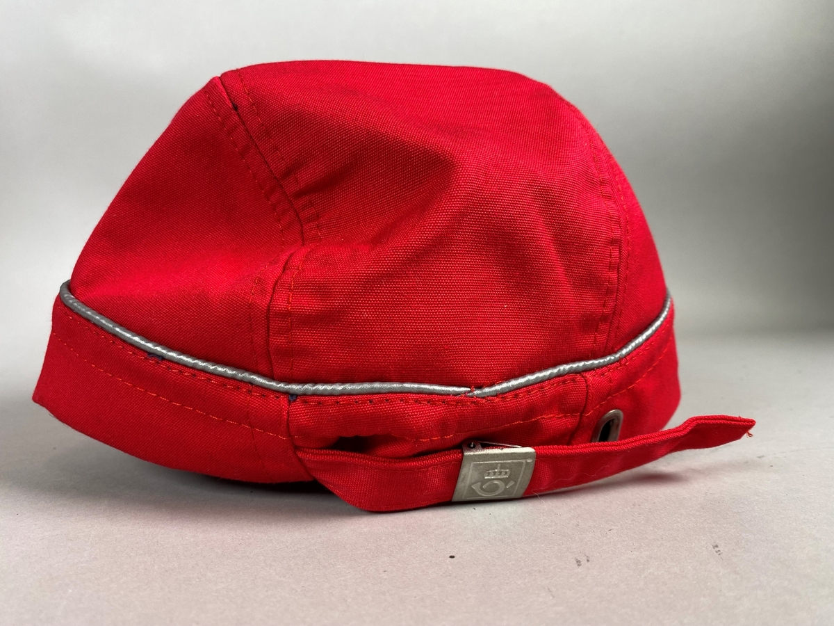 Rød caps med solskygge og Postens logomerke. Capsen har et smalt refleksbånd som går rundt, og hempe og bånd for justering av størrelse på baksiden.