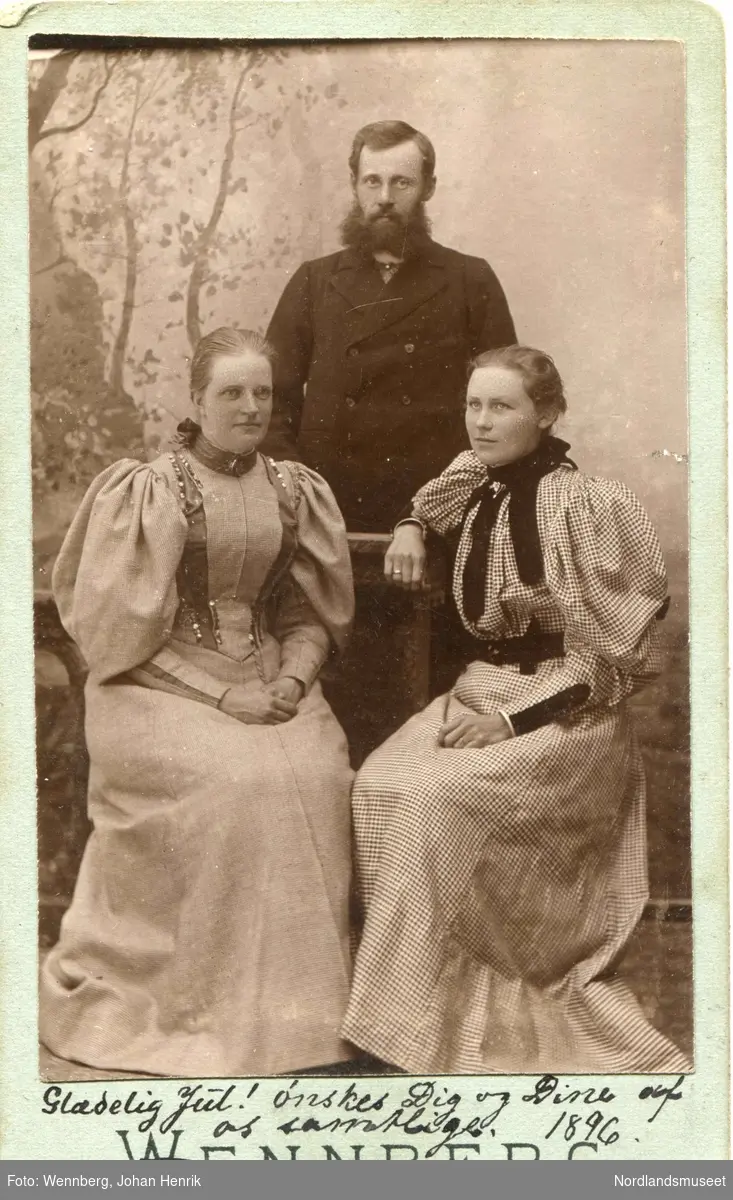 Gruppebilde. En mann står bak to kvinner foran et malt lerret. Fotoatelier. Severin og Sigrid Tresseng og Frøken Trønnes.