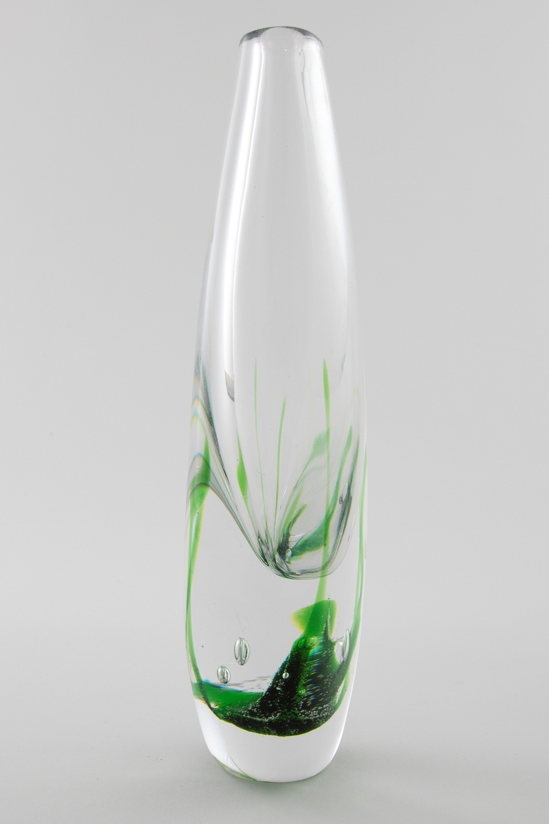 Flattrykket dråpeformet vase i klart glass med liten sirkulær munning. Dekor i grønt i underfangsteknikk, som minner om alger som vokser opp fra vasens bunn. En del luftbobler i glassmassen.