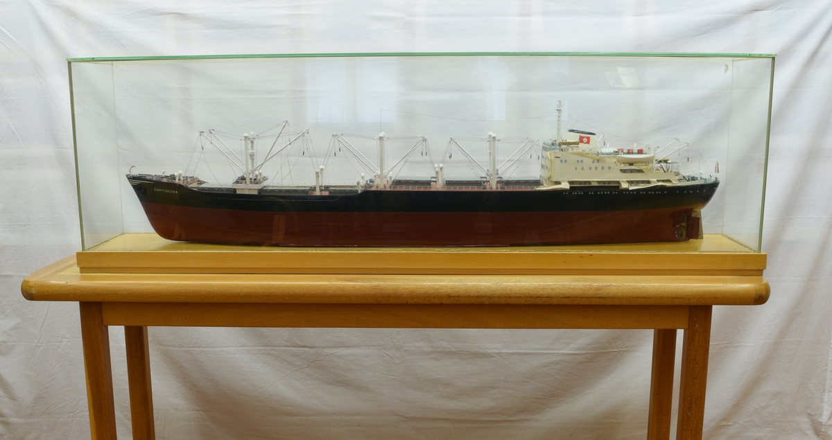 Båtmodell med glasslokk i som kan løftes av plate med påmontert modell. Plate og listverk i lakkert lyst tre. Modell i tre, plast og metall.