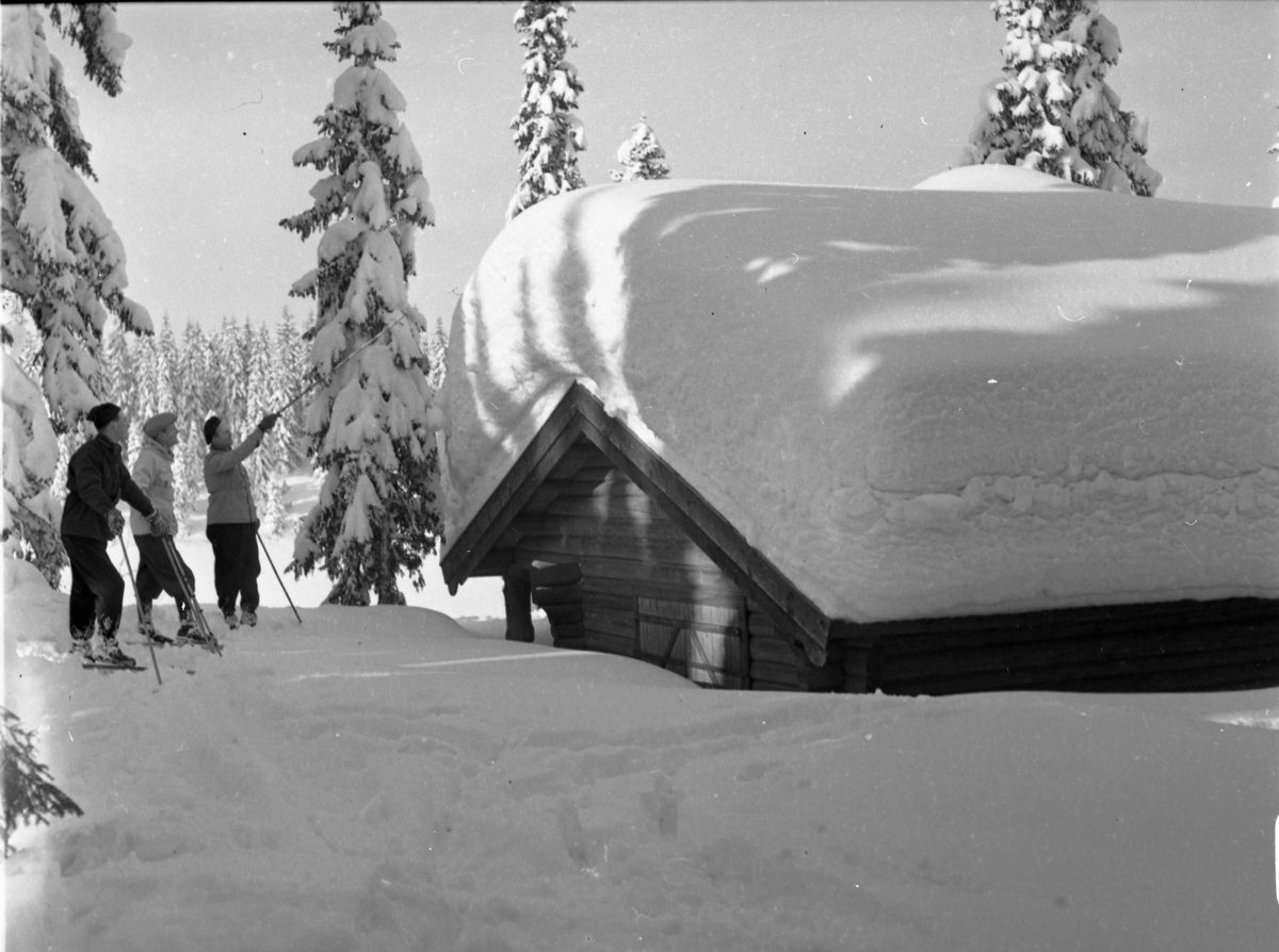 Eksteriør av snøkledd hytte. Tre vinterkledde menn på ski står ved siden av hytten.
