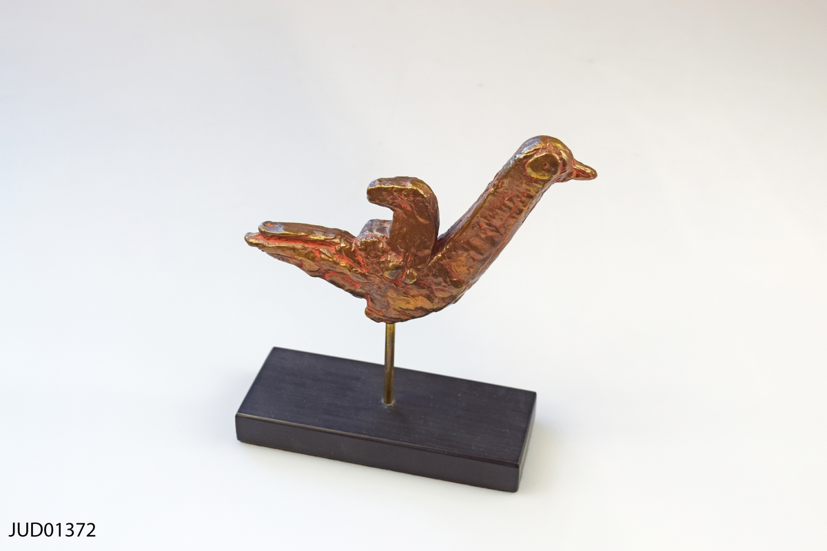 Fågel med hundfigur på ryggen i brons. Monterad på pinne i rektangulär bas i trä. 
Titel: "Fågel med hundvalp"