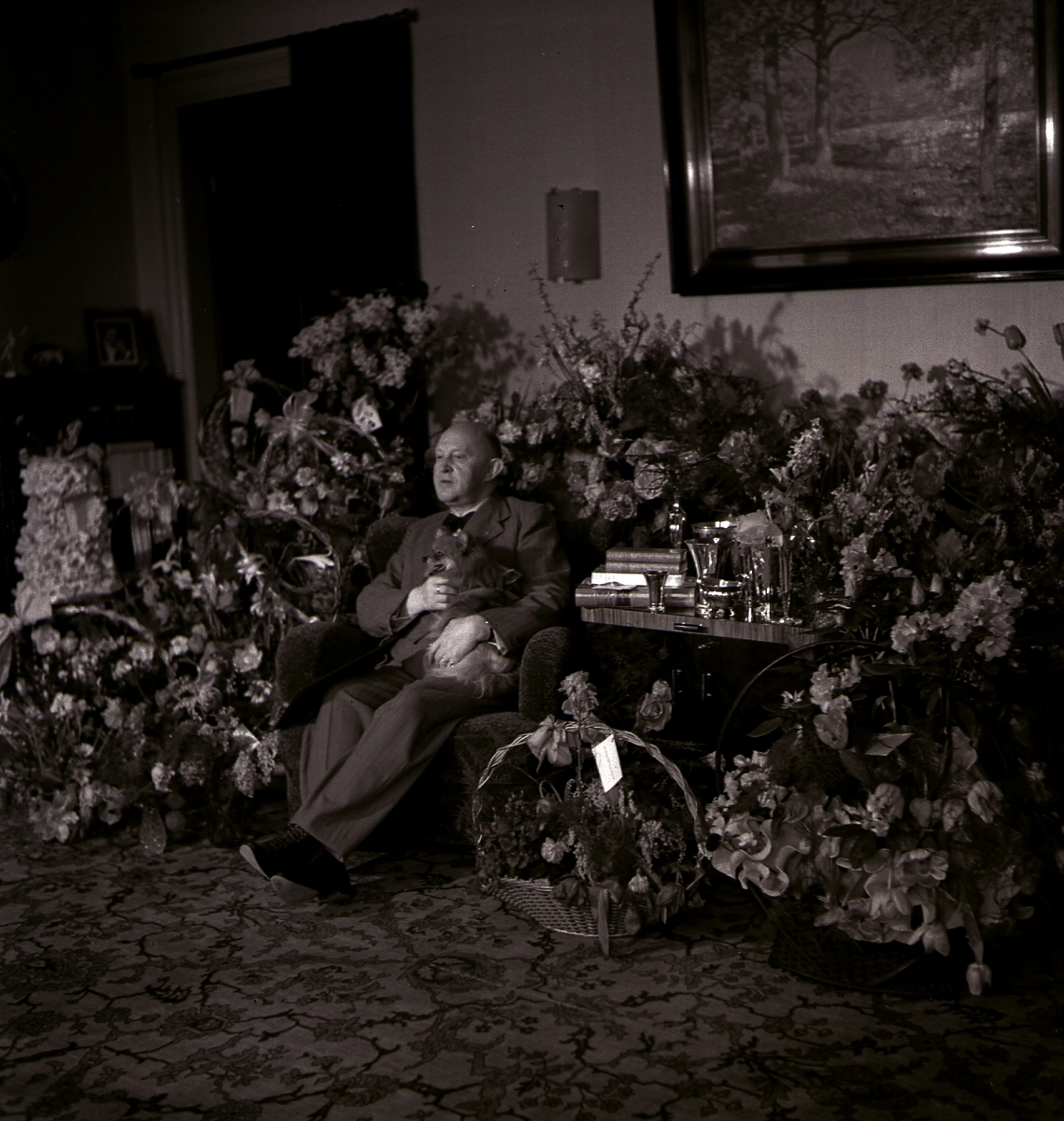 Köpman Edvard Andersson, innehavare av Bröderna Anderssons pappershandel, sitter i sitt vardagsrum med en liten hund i knäet, omgiven av mängder med blommor på sin 60-årsdag.