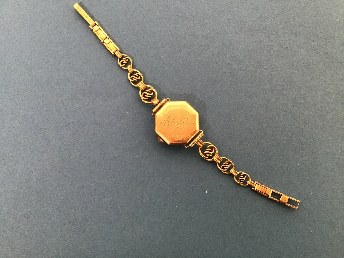 Armbandsur, dameur, i metall, urskiva er kvit med arabiske tegn for markering av 5-minuttsintervall, stigande frå 1 - 12. Metallreim. Sjølve urkassa er åttekanta - oktogon.
