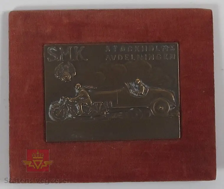 Plakett laget av metall i en bronse-brun farge, rammet av rødt velour stoff. På plaketten er det utformet en bil og motorsykkel som tilsynelattende kjører om kapp.