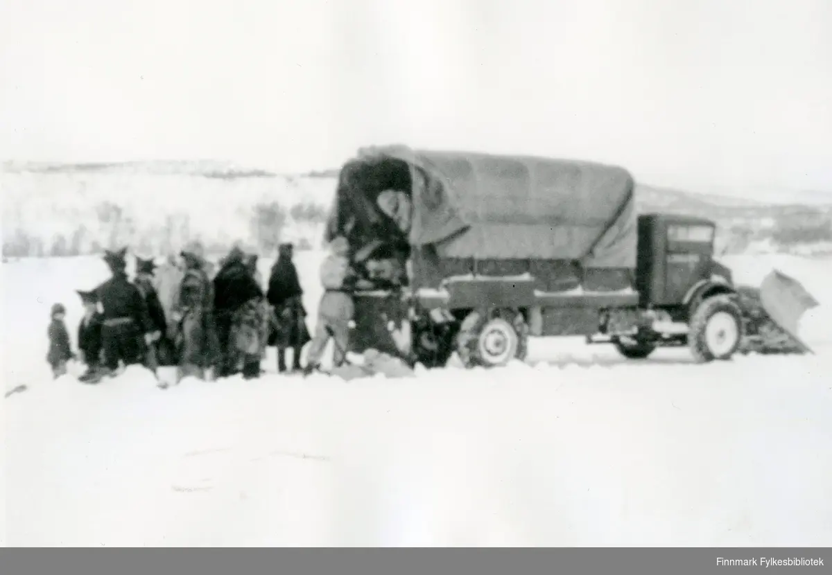 Militært kjøretøy med snøplog på isveien. Samer i pesk med stjerneluer og soldater i hvite vinterdrakter er fotografert ved bilen. Bildet er tatt i 1945, ref. Norsk veimuseum.