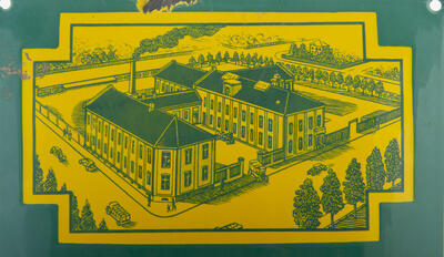 Reklameskilt for Norrøna skofabrikk i grønn- og gullakkert metall. Skiltet viser en tegning av fabrikklokalet.
