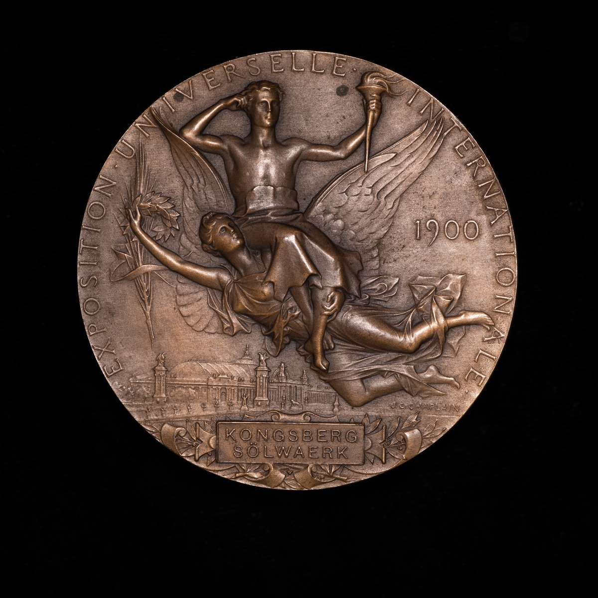 Bronsemedalje fra utstilling i Paris 1900. På advers kvinnehode i profil mot høyre. På revers mann med fakkel i venstre hånd sitter på ryggen til flyvende kvinnelig engel.