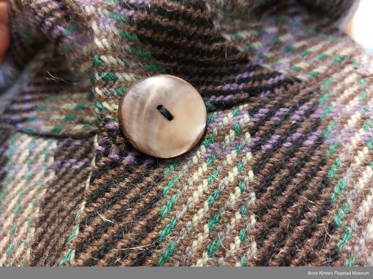 Ullkåpe i kypert vevet stoff  med ulike farger i striper. Spesiell lomme med "klaffen" som en forlengelse av en fold fra halsringningen. 2 knapper i halslinningen, den ene festes gjennom en åpning i sømmen til halslinningen.
