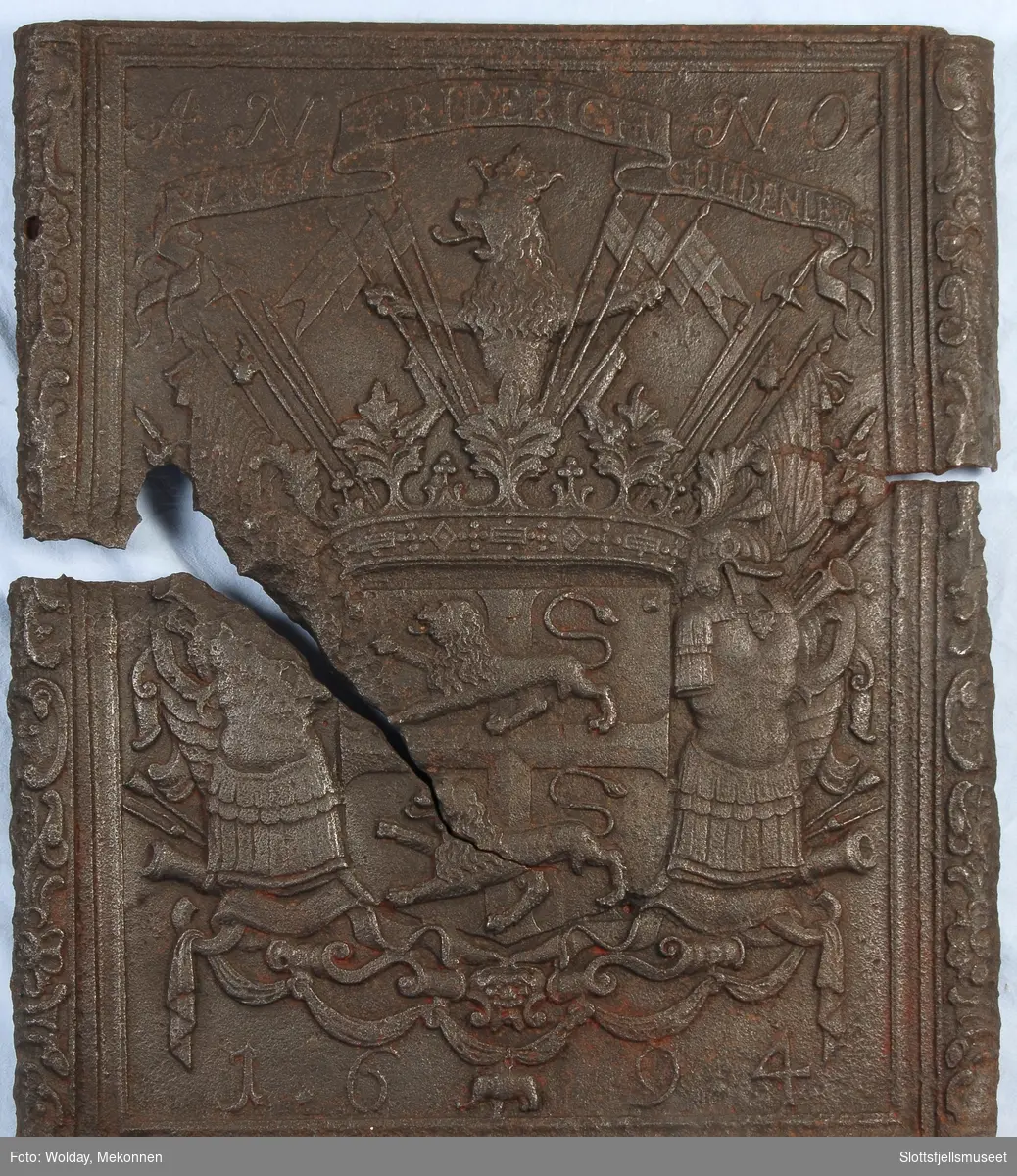 Gyldenløves våpern, omgitt av krigerske emblemer, derover på knekket bånd "ULRICH FRIDERICH"
nederst 1694