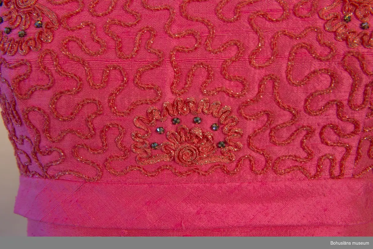 Rosa långklänning band något över midjan som löper runt klänningen. Över bandet, på klänningens framsida, mönstrat tyg ornamenterat med musslor i oregelbundet mönster. Baksidan har ett släp fastsytt i bandet.
