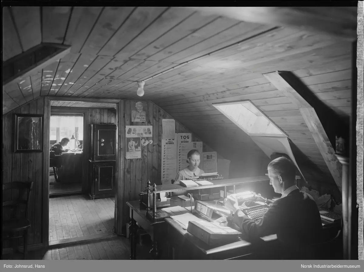 Kontor for Kaare Østdahl med kontorarbeidere i arbeid. En kvinne og en mann skriver på skrivemaskin, en annen lener seg over bøker på skrivebord. Safe stående i ett av rommene.