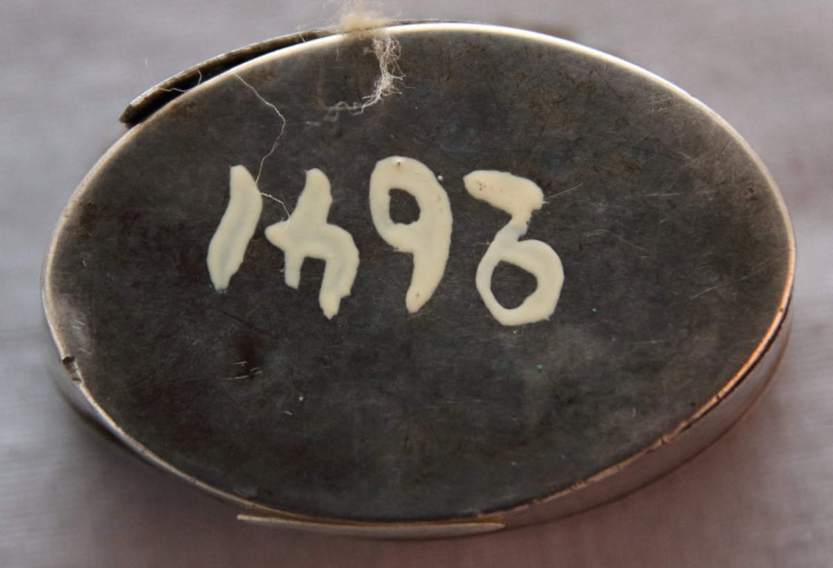 Oval dosa av silver. Med ett lock med gångjärn. Ytterkanten på ovansidan av locket har en cisilerad bård av stiliserade blad. I mitten av locket står initialerna "B. S. D. " och "G. A. S." . Asken är stämplad på insidan av botten med bokstäverna "MOB" (Michael Olof Barkman,1813-1851, Varberg) och med "N4" (år 1843) samt två svårtydda stämplar.
