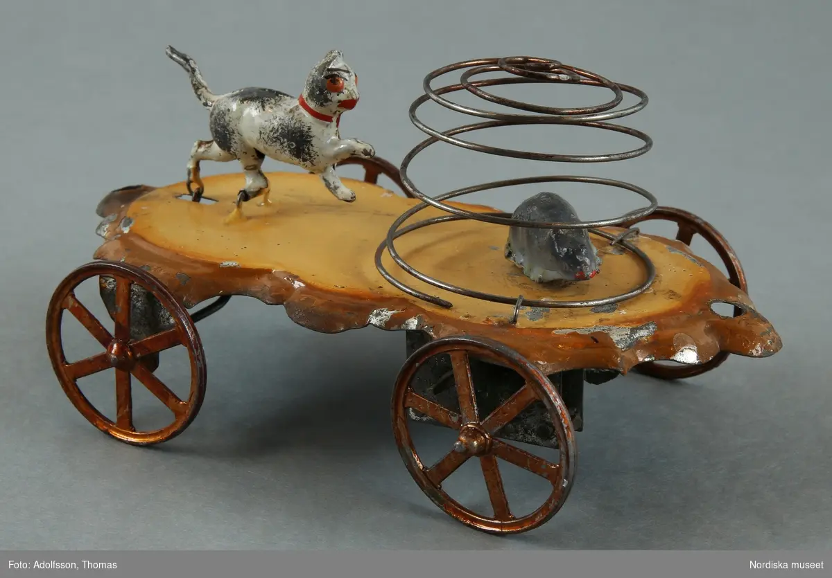 Inventering Sesam 1996-1999:
H 8   L 15,5   B 9 (cm)
Katt, leksak, och råtta på vagn, av lackerad plåt; vagn med fyra hjul lackerade för att föreställa koppar, lackerad gulbrun med mörkare bruna konturerade kanter. På vagnen en katt, vitlackerad, med rött halsband, och en grå mus i en bur i, formad likt en spiral av ståltråd. När vagnen rullas framåt rör sig katten medan musen snurrar runt i buren.
Har tillhört Albert Harling, född 1879. Fadern tullförvaltare i Norrköping.
ELB / 70 /=Eva Lis Bjurman/
Charlotta Dobson Hoffman jan 1998