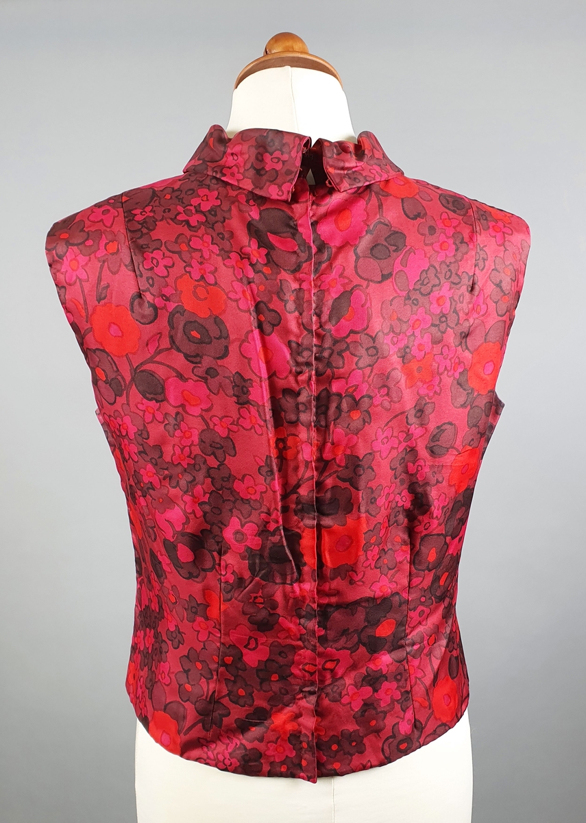Rød bluse av blomstermønstret silke. Blusen er uten ermer og med stående krage. Glidelås i ryggen. I fallen nederst er det innsyd lenke med blylodd.
