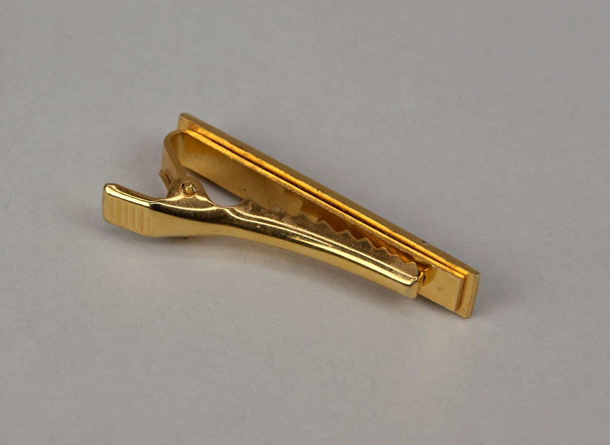 Slipsklämma av guldfärgad metall. Rektangulär form indelad i sex fält, i det sista finns SJ:s logga. Fästs i slipsen med räfflad klämma.