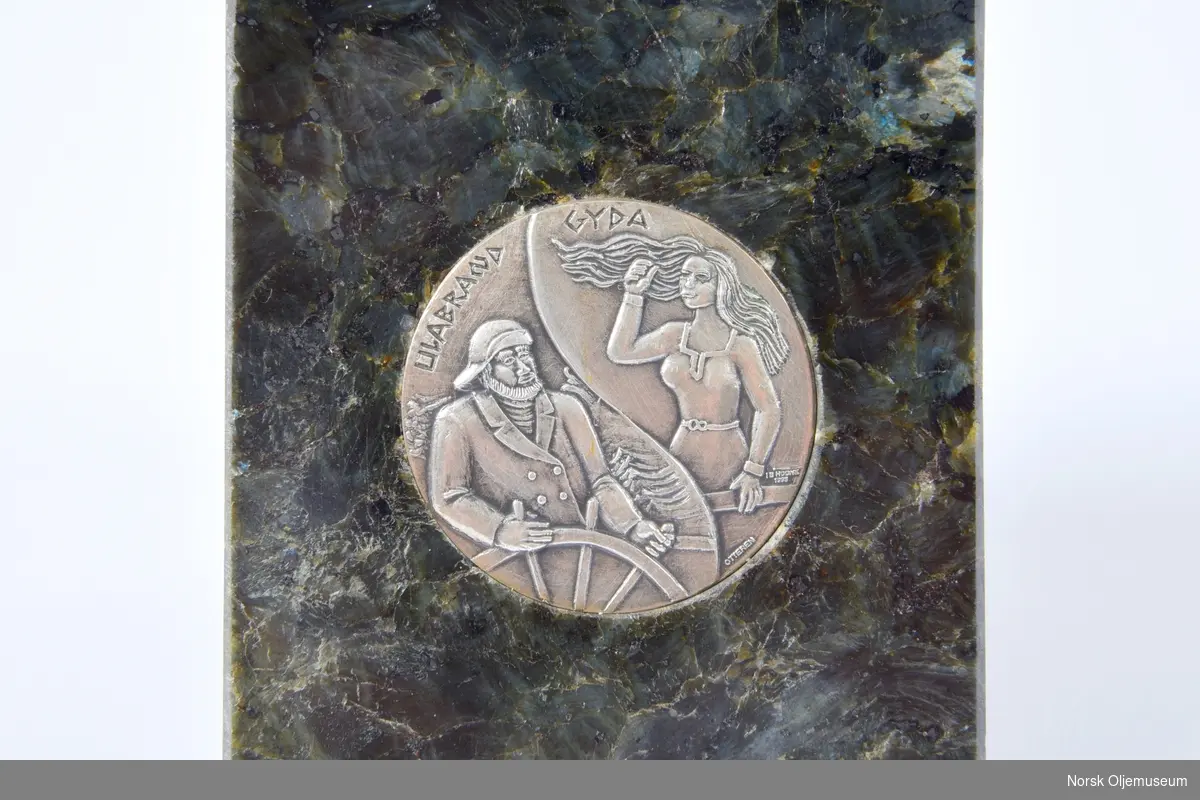 Tilskåret steinemne med innfelt relief i metall. Reliefet viser en sjømann, og en kvinne med flagrende hår. Teksten identifiserer dem som Ulabrand og Gyda.