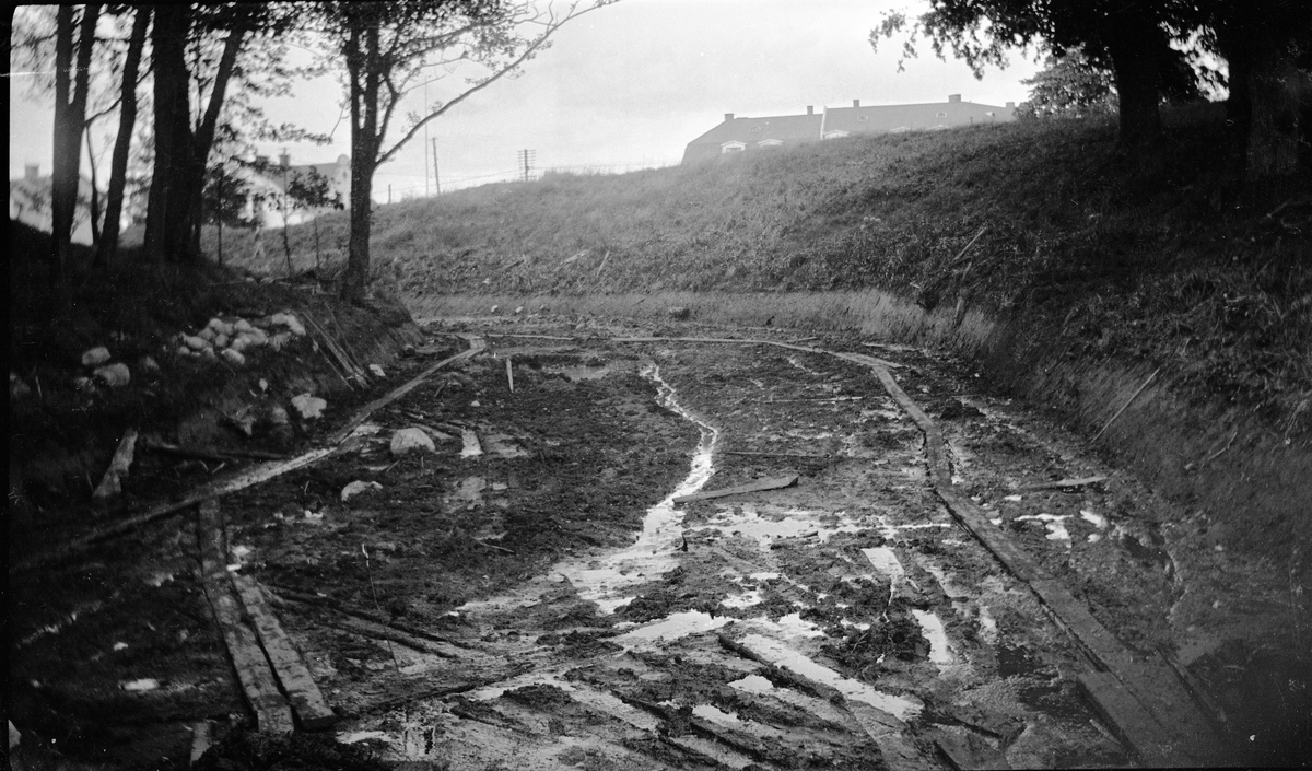 Den inre vallgraven vid Rumlaborg i Huskvarna är färdigutgrävd i augusti 1932. Vid undersökningen av vallgraven påträffades bl.a. timmerrester och en halv stockbåt med medeltida datering. Fotot togs i samband med arkeologiska undersökningar vid borgen och undersökningen av vallgraven blev möjlig med hjälp av en elektrisk pump som pumpade bort vatten, samt tack vare att en spåntvägg hade uppförts tvärs över vallgraven mot Huskvarnaån. Föregående foto visar samma vy efter att spåntväggen tagits ner och pumpen slutat gå.