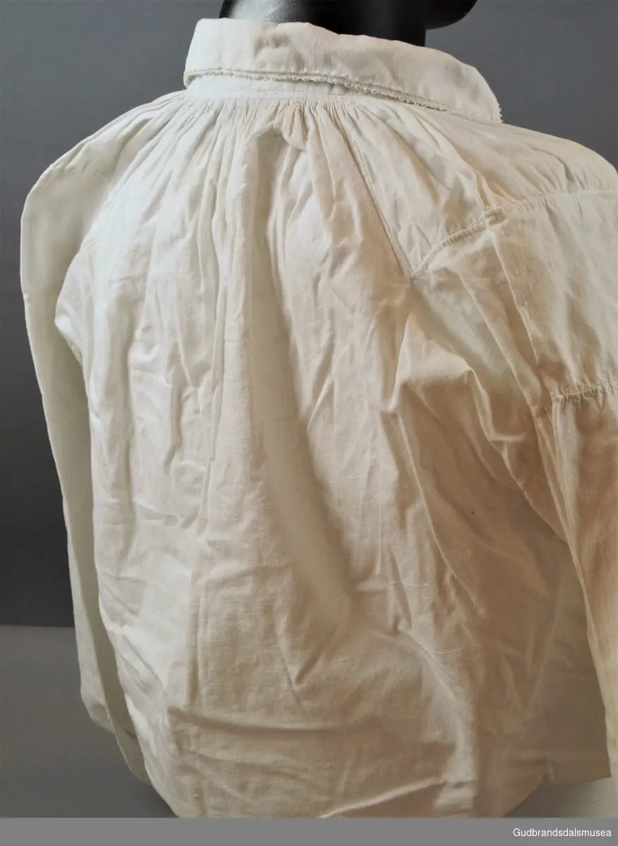 Kvitt bomullsskjorte med "kjæ'såm" og hullagger (halvløkker) langs kragebord og ermlinning som i tillegg har kryssesting m/slyng. Knapphull på erme- og halslinning. Halssplitt som er sydd med 3 cm. lang "kjæ'såm" i enden.
