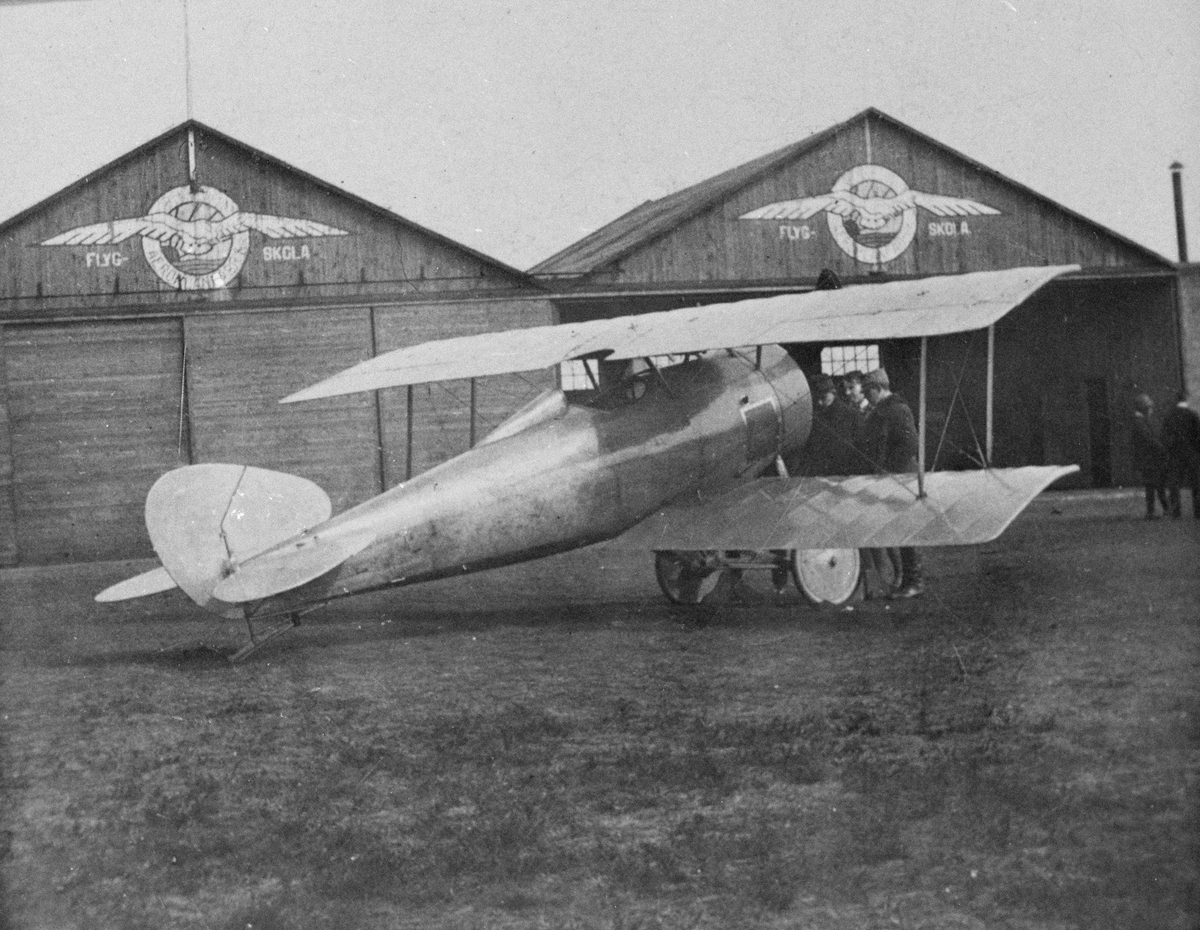 Flygplan Thulin N står framför Thulins flygskola i Ljungbyhed, cirka 1917. Tre män står vid flygplanet. Vy snett bakifrån.