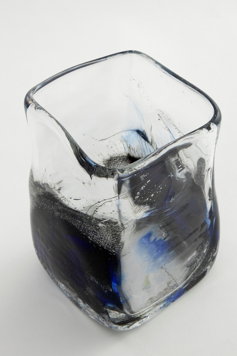 Kvadratisk vase i klart glass med tykk bunn. Nedre del er delvis utbulet, og dekorert med metallnett, luftbobler og koboltblå partier. Øvre del er utført i klart glass med enkelte luftbobler.