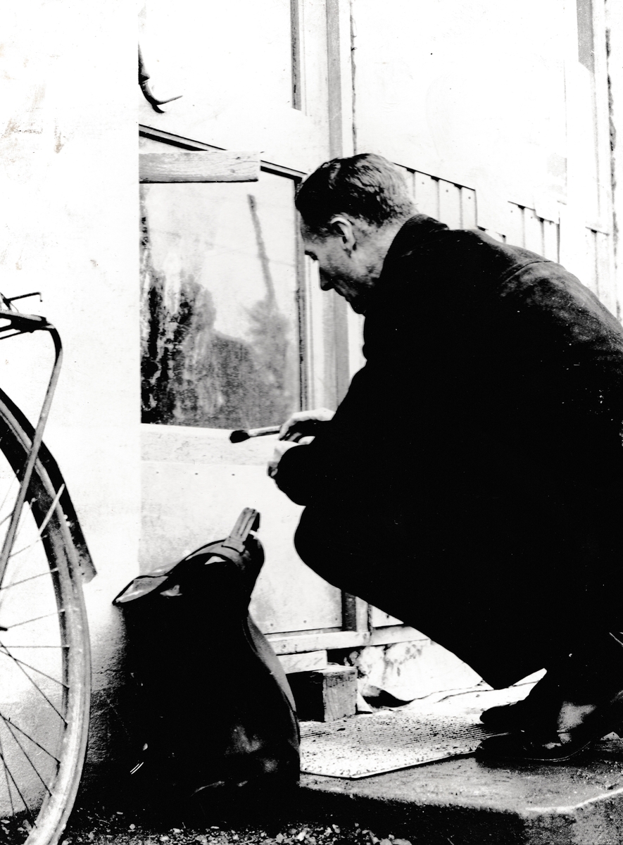 En mann sitter på huk foran en glassrute med en pensel i hånden. Veske, sykkel.