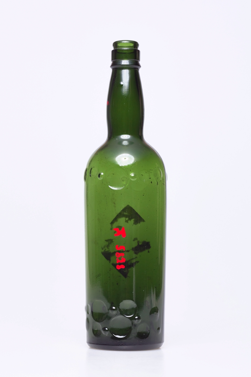 Grønt glass, fabrikklaget. Det er to rekker med påstøpte merker på flasken og rester etter merkelapper.