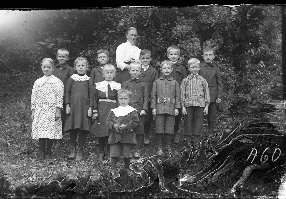 Lärarinnan Bertha Engström (fotografens mor) med en skolklass om 12 barn från småskolan i Sällstorp, samlade utomhus. Fr v: Elin (syster till Horse-Herman),  Kulla-Gunnar (Ulvatorp f. 1906), Sofia (Sves i Holmåkra), Karl Johansson (Sväs-Karl Ulvatorp f. 1907), Lelle Hansa-Oskar (f. 1907) och längst till höger står Gustav J. (bror till Alice i Larsa) och framför honom i randig skjorta Gade-John (Ulvatorp f. 1906), övriga är okända. Meddela oss gärna om du vet namnen på någon! (Negativet har skador nedtill)