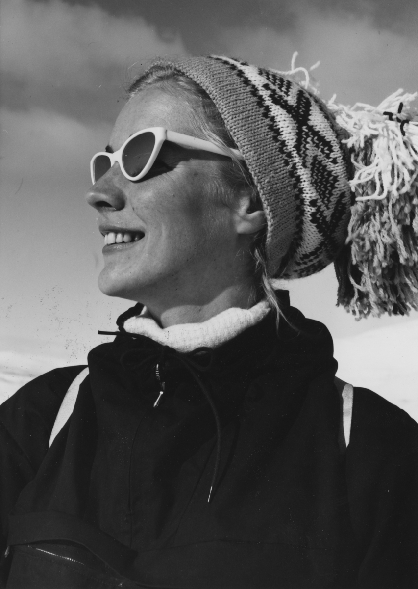 Portrett av en kvinnelig modell på tur en solfylt vinterdag. Hun har klassisk strikkelue med stor dusk og solbriller.