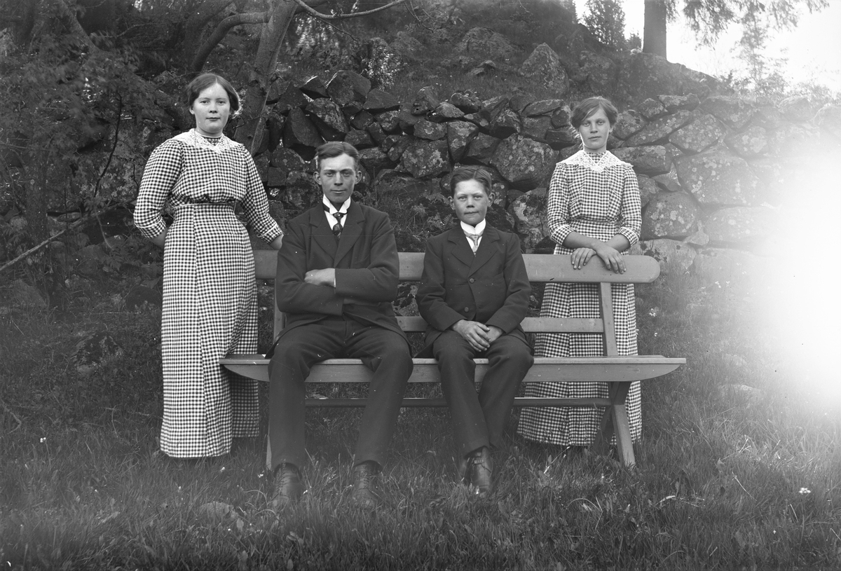 De fyra äldsta syskonen Eliasson på Svärtingskulla, Ås socken, samlade utomhus vid en hög stengärdsgård. Bröderna sitter på parkbänken och döttrarna står i likadana klänningar på var sida om bröderna.
(Se även bilderna GEA010, GEA021, GEA022)