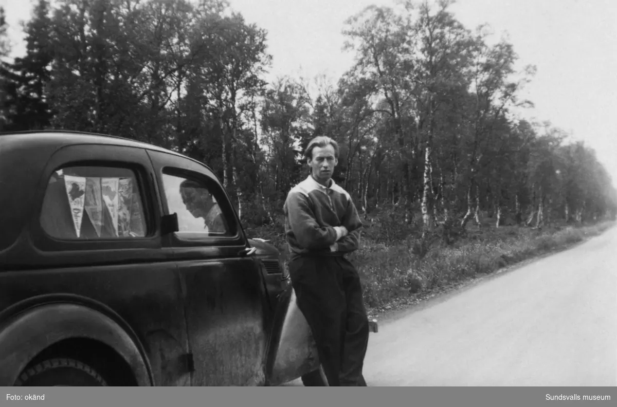 En ung man står lutad mot sin bil invid landsvägen. I bakrutan hänger vimplar från tidigare resmål. På bildens baksida står: Sten Friberg med sin bil Ford Eifel 1938. Kortet taget senare delen av 1940-talet. Ur en samling från Byn, Norrbäcken, Sättna.