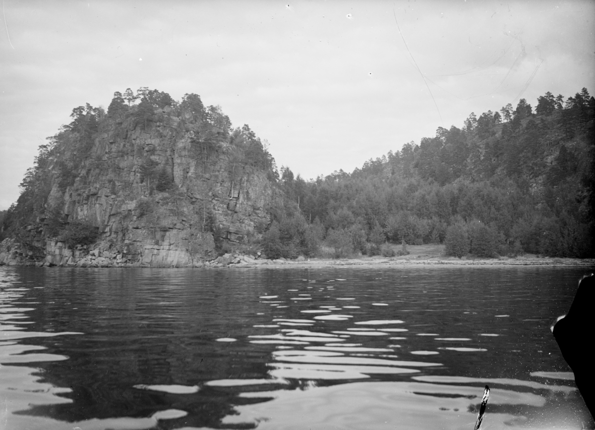 Landskapsbilde av fjellknauser med skog, langs vannet. Tekst fra Teigens katalog: "Svelviks Historie, Svelviks museum etc. se også: Drammens Tidende, se også: Hurum Bygdebok 4938"