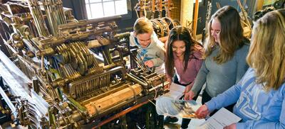 Bildet viser fire unge jenter som holder i penn og papir  ved siden av en maskin.. Foto/Photo