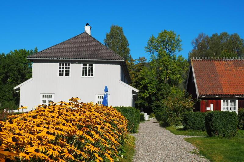 Bildet viser en lys bygning i tre og to etasjer og et lavere, rødt uthus. I forgrunnen er det et gedigent bedd med gule blomster