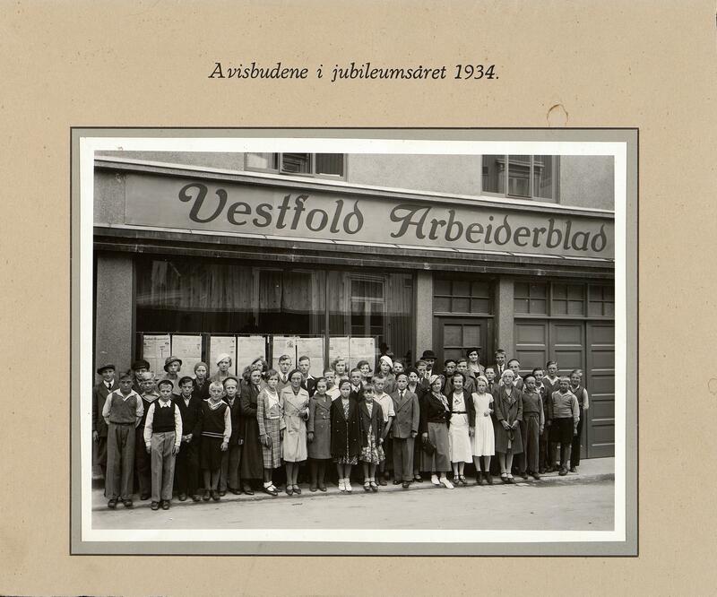 Et sort-hvitt foto er limt på en papplate. Bildet viser en gruppe mennesker som står oppstilt foran et bygg med skriften Vestfold Arbeiderblad på veggen.