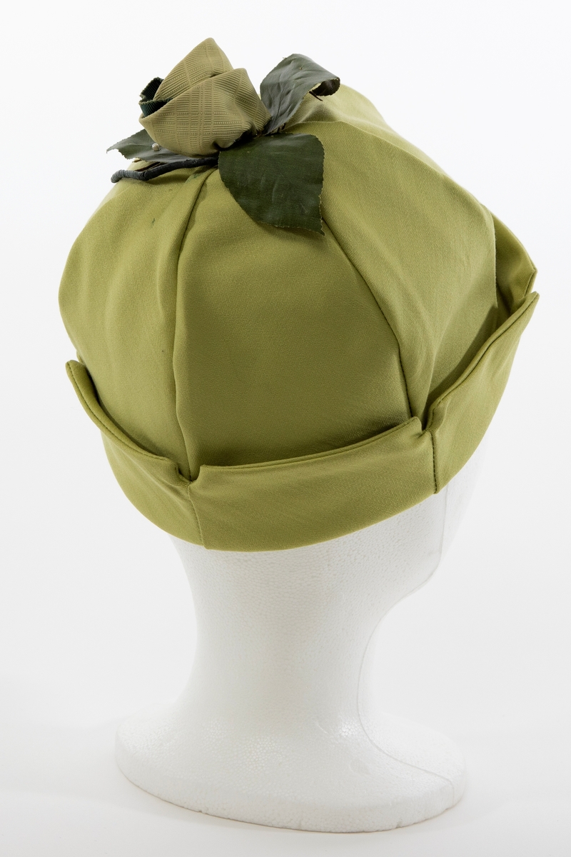 Klokkeformet, sommerlig hatt med opprett nederst. Satengvevd, halvblankt stoff. Hatten er dobbel, dvs at den er sammensydd av 2 x 6 sektorer, slik at den har lik konstruksjon innvendig som utvendig. Hatten skal være oppbrettet, og det er korte splitter i nederkant mellom hver sektor. Oppbrettet gir det en takket kant på oppbretten. På toppen av hatten er det påsydd en tekstilrosett, en slags "Dior-rose", konstruert av et limefarget syntetstoff med stripet og rutet overflatestruktur, omkring et innvendig, mørk grønt, ripsartet silkebånd. Mellom rosen og hatten er det festet 3 mørkegrønne blader av sterkt appretert tekstil, med stilker av wirer og én tykkere, vokset hovedstilk. Det erne bladet er prydet med en påsydd, ørliten. perlemorskimrende perle. Det kan se ut som også de 2 andre bladene har hatt en liten perle.