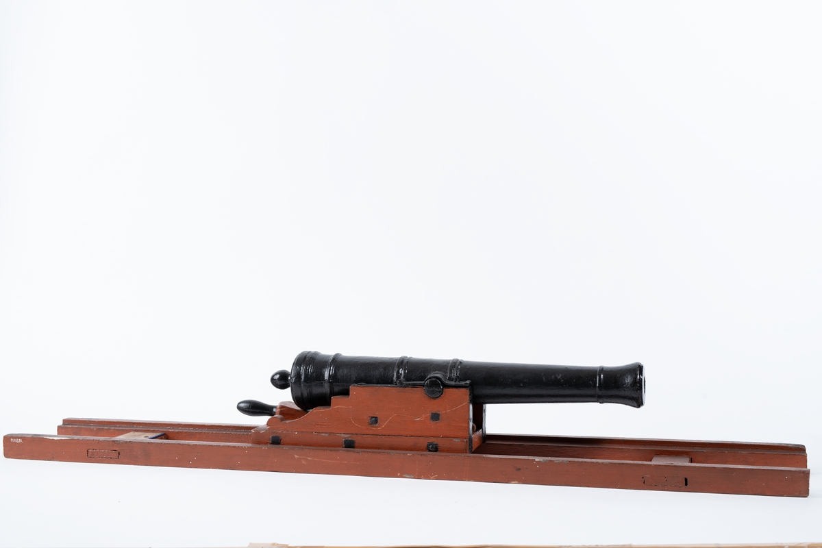 Kanonmodell: galérlåda med släde, beslagen, samt kanon. Lavetten av trä, rödmålad. Kanonen av järn, svartmålad. Tillbehör: 1 st ställhult. Lavetten L = 955 mm B = 142 mm H = 130 mm.