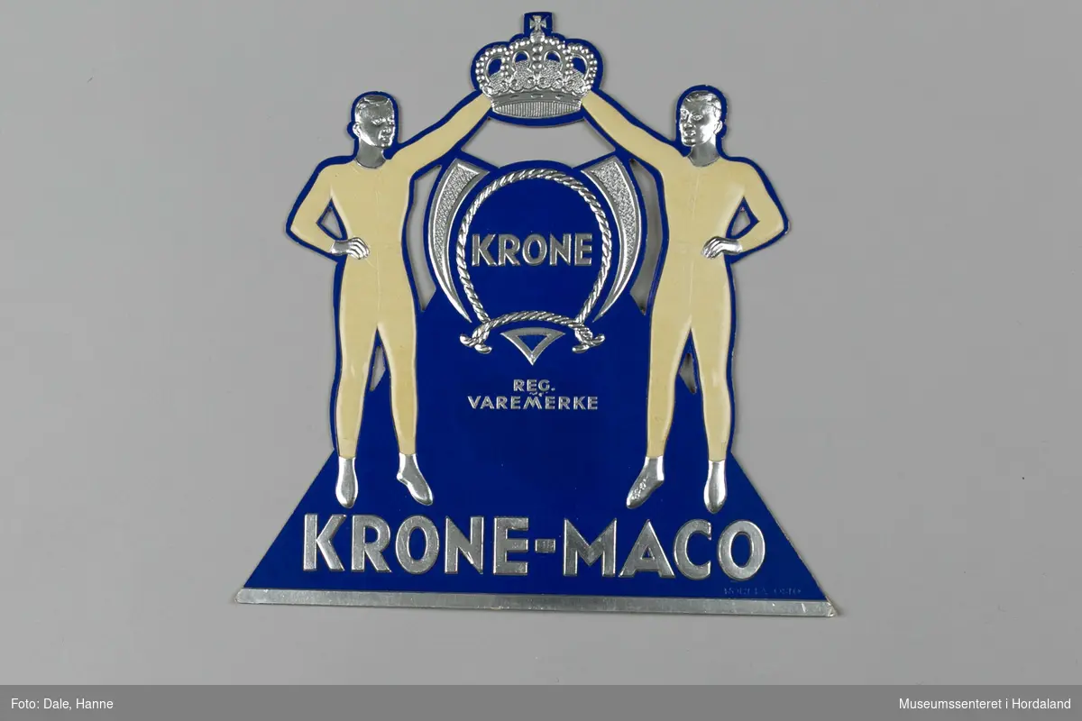 Reklame for Krone-Maco og Krone-merket frå Salhus Tricotagefabrik ved Bergen. Den er laga i påtrykt papp med "fot" på baksida slik at den kan stå på ein disk eller i ei hylle.