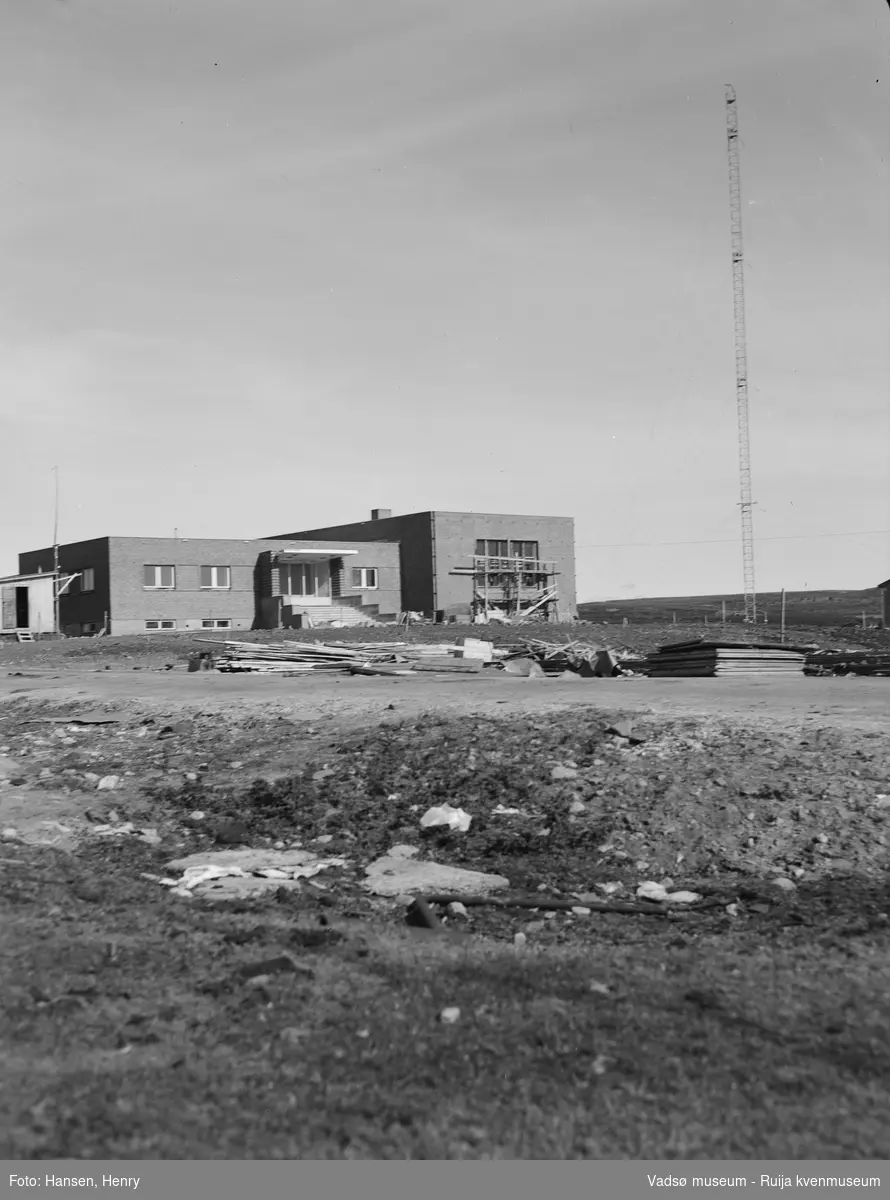 Det nye NRK-bygget i Vadsø ble reist 1948-49. Bygningen er ennå ikke helt ferdigstilt og vi kan vi se stillaser,  forskalingsmateriale, grushauger og arbeidsbrakke på og rundt bygningen. Bak bygget ser vi en av de 2 radiomastene.