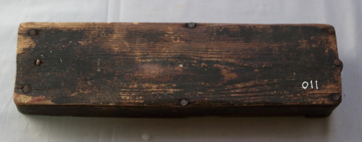 Rektangulär ask i trä med en avancerad låsordning inbyggd i locket. Botten och sidorna är spikad med metallspikar. Träet är mörkt lackat. I skrinet finns en liten 7 cm lång rakborste.