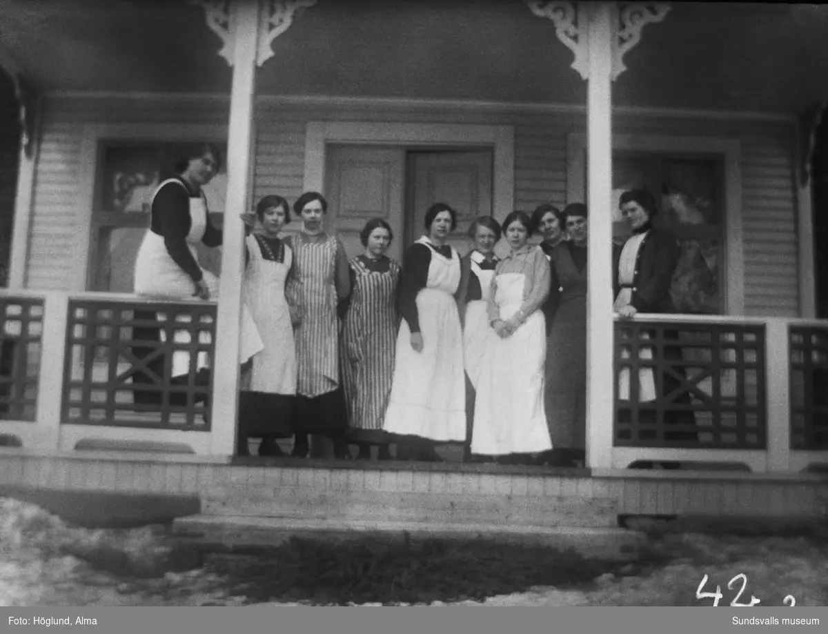 Gruppbild på en veranda med tio unga kvinnor, alla med förkläden