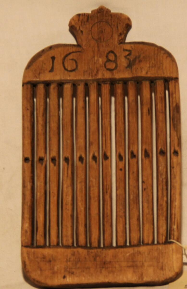 Bandvävsked av trä, med profilerat överstycke på ena sidan inskuret "1683", på den andra "K I D". 12 tinnar med hål i mitten. - Höjd 21 cm. Bredd 11,2 cm. - Gåva av Jonas Näslund.