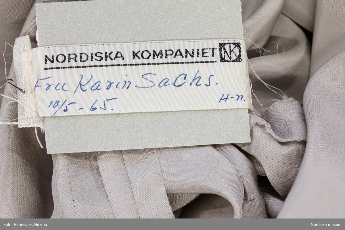 Klänning monterad på docka. Beställd av Karin Ekelund Sachs (1913-1976) firad skådespelerska på 1940-talet och Radioteaterns första kvinnliga producent 1961. Hon var gift med NK-direktören Ragnar Sachs. och stamkund i varuhuset i decennier, hon kallades "NK:s första dam". Aftonklänningen av silverlamé med drapering över ena axeln syddes upp inför en middag på Kungliga slottet och levererades i maj 1965. Etiketter i plagget.